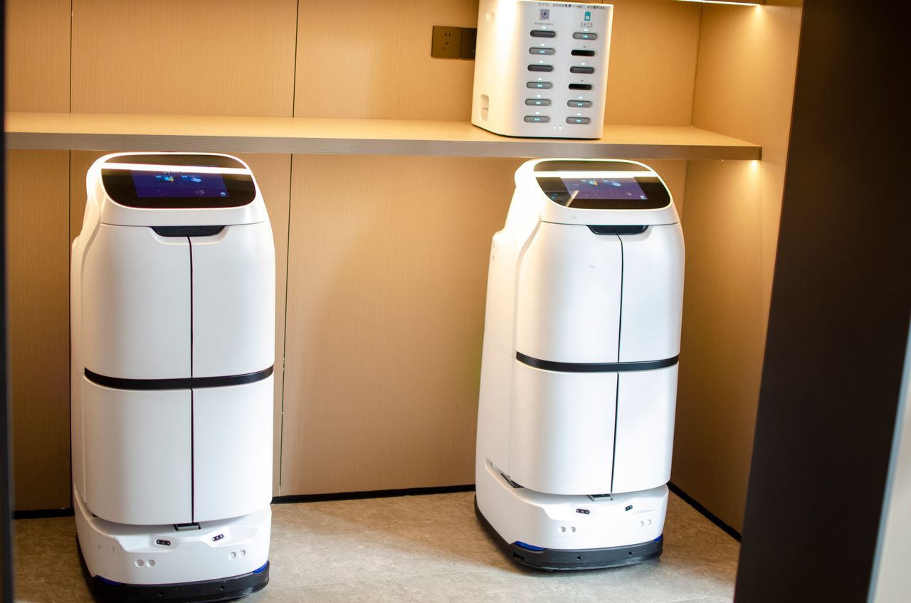 services-02 Two white robotic delivery systems located in a modern indoor room with soft lighting.